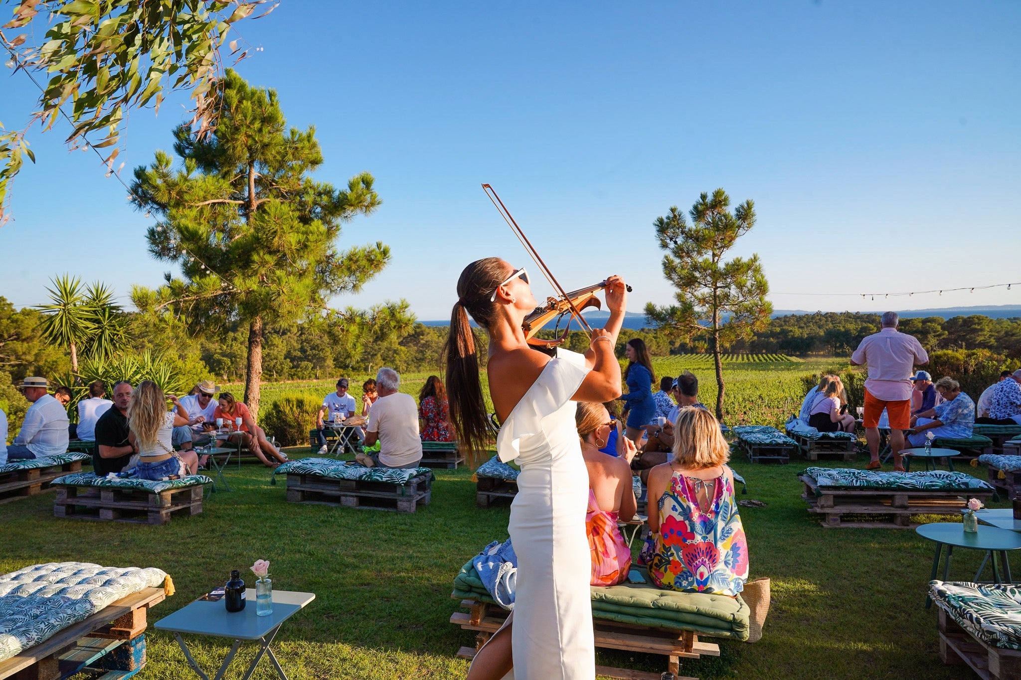 Une violoniste joue de la musique live pour un public assis lors d'un événement oenotourisme au Domaine de la Sanglière à Bormes-les-Mimosas, en Provence. Notre vignoble, certifié HVE3 et AOP Côtes de Provence La Londe, offre une vue panoramique imprenable sur la mer Méditerranée et son terroir de schiste. Un moment de partage et de dégustation inoubliable pour la famille.